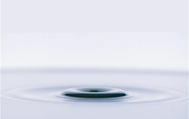 Close-up of a droplet creating ripples in a calm, pale blue liquid surface with a central swirling pattern.